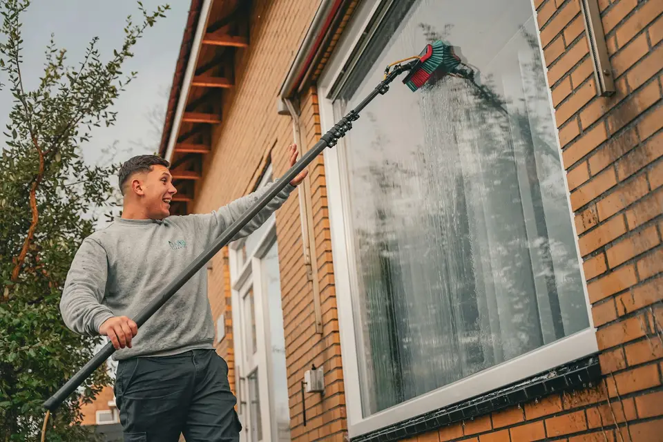 Een glazenwasser maakt met een lange steel een groot raam in een bakstenen gevel schoon